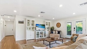 A spacious living room with a large sofa, a coffee table, and a television. There are cabinets with glass doors and a large window. - Valley & Vine- Pool- Walking Distance to Old Town! (Scottsdale)
