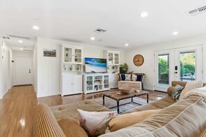 Ein geräumiges Wohnzimmer mit einer großen Couch, einem Couchtisch und einem Fernseher. Es gibt Schränke mit Glastüren und ein großes Fenster.
