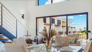 Un espace de salle à manger moderne avec une table en verre, des chaises blanches et un vase décoratif. Un escalier est visible sur la gauche, et un balcon avec vue sur les bâtiments se trouve à l’arrière-plan.