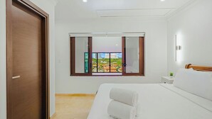 Une chambre d’hôtel avec un grand lit, une fenêtre donnant sur un paysage urbain, une porte en bois et une petite table de chevet agrémentée d’une plante.