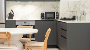 Eine moderne Küche mit dunklen Schränken, einer Mikrowelle und einer Kaffeemaschine. Es gibt einen Essbereich mit einem weißen Tisch und zwei Stühlen.