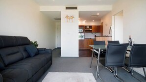 A living room with a black sofa, a dining table with chairs, a kitchen in the background, and a white pillar with a giraffe picture. - CBD Apartment - Walk to Shops & Attractions (Launceston)