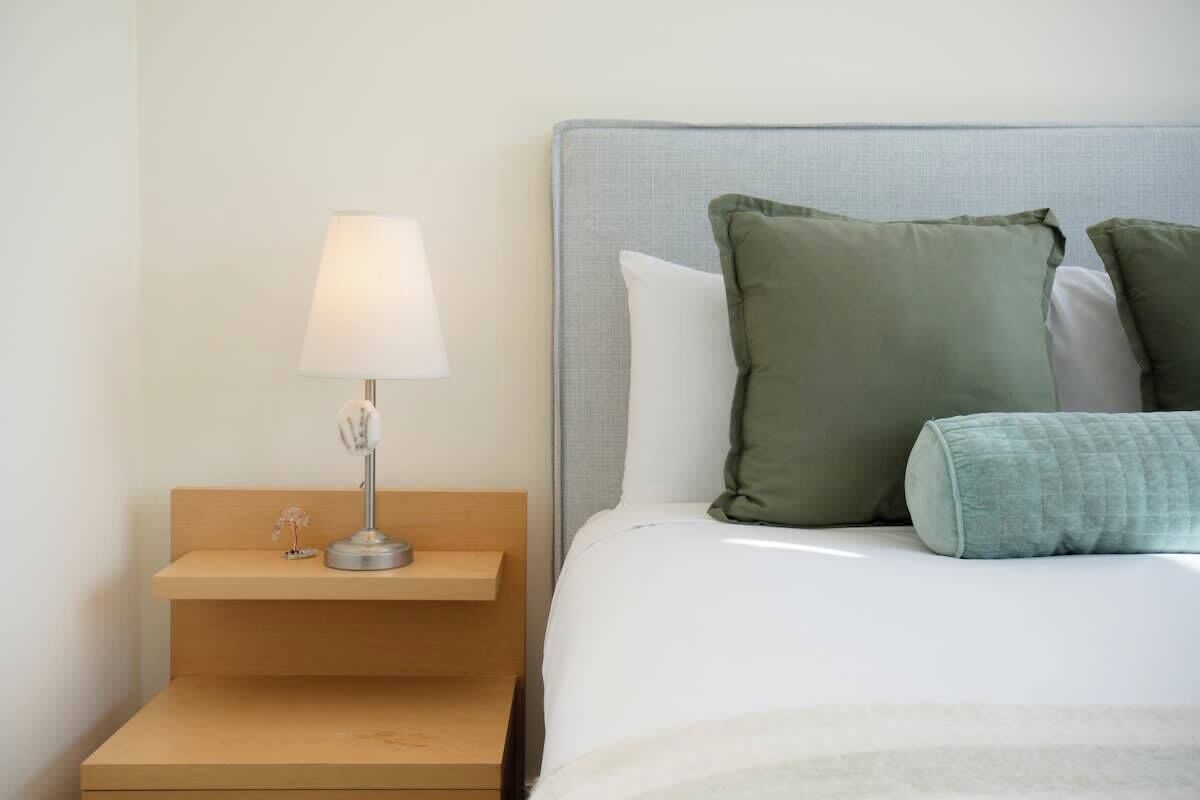 Una cama bien tendida con almohadas, una lámpara de noche y una mesita de madera.