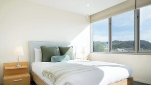 A bedroom with a large bed, a nightstand, a lamp, and a window with a view of hills. - CBD Apartment - Walk to Shops & Attractions (Launceston)