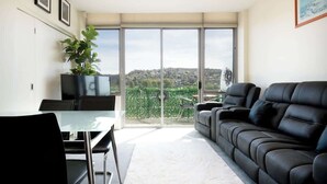 A modern living room with a black leather sofa, a glass dining table, and chairs. There is a large window with a view of a green landscape. - CBD Apartment - Walk to Shops & Attractions (Launceston)