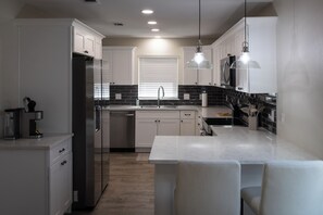 Uma cozinha moderna com armários brancos, geladeira de aço inoxidável, ilha central com banquetas e iluminação pendente.