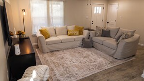 Una sala de estar con un sofá seccional color beige, un tapete estampado, una pequeña mesita auxiliar y un ventilador de techo.