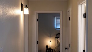 Un pasillo con piso de madera, paredes blancas y una puerta que da a un dormitorio con cama y lámpara.