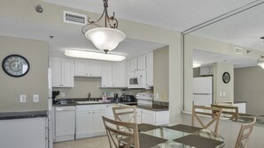 Une cuisine moderne avec des meubles blancs, un coin repas doté d’une table à plateau en verre et de chaises en bois, et une horloge bien visible au mur.