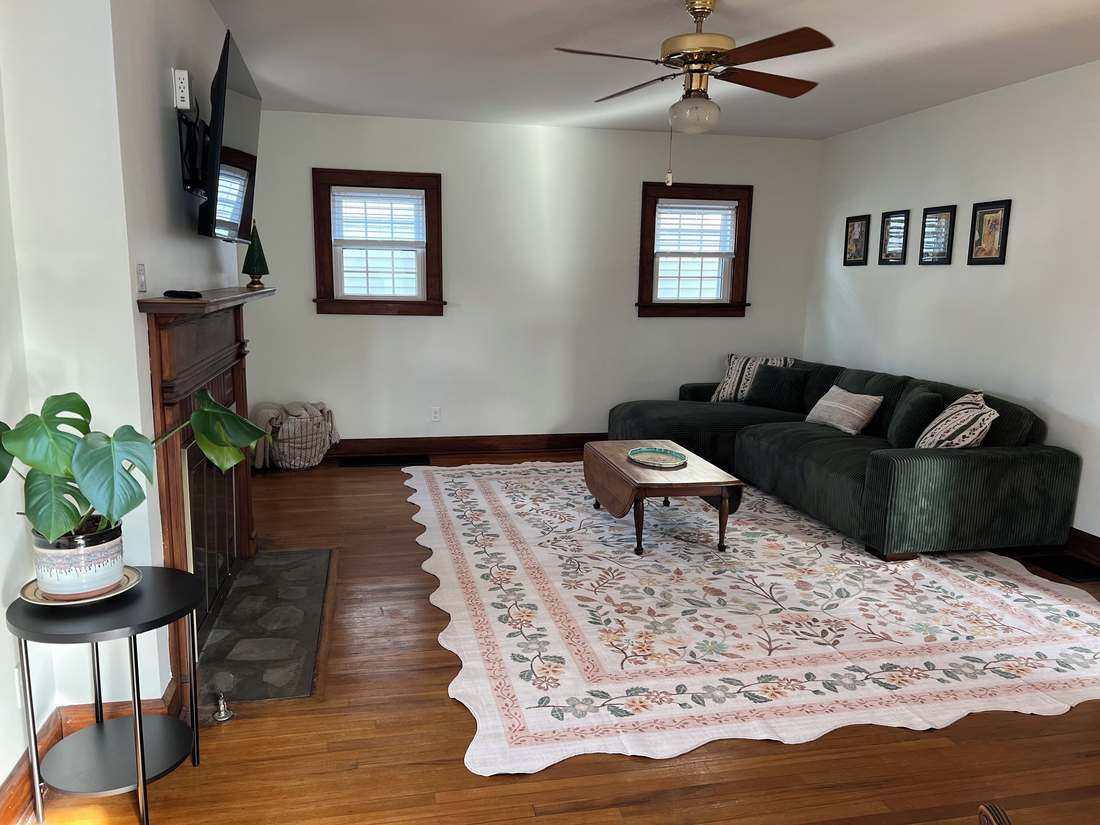 Ein Wohnzimmer mit einem dunkelgrünen Sofa, einem Holztisch, einem Teppich mit Blumenmuster und einem Deckenventilator.