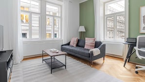 Ein modernes Wohnzimmer mit einem grauen Sofa, einem kleinen Couchtisch und einer Lampe. Große Fenster sorgen für natürliches Licht und bieten Blick auf Gebäude.