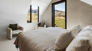 Een slaapkamer met een groot bed, twee fauteuils en uitzicht op een bergachtig landschap door een groot raam.