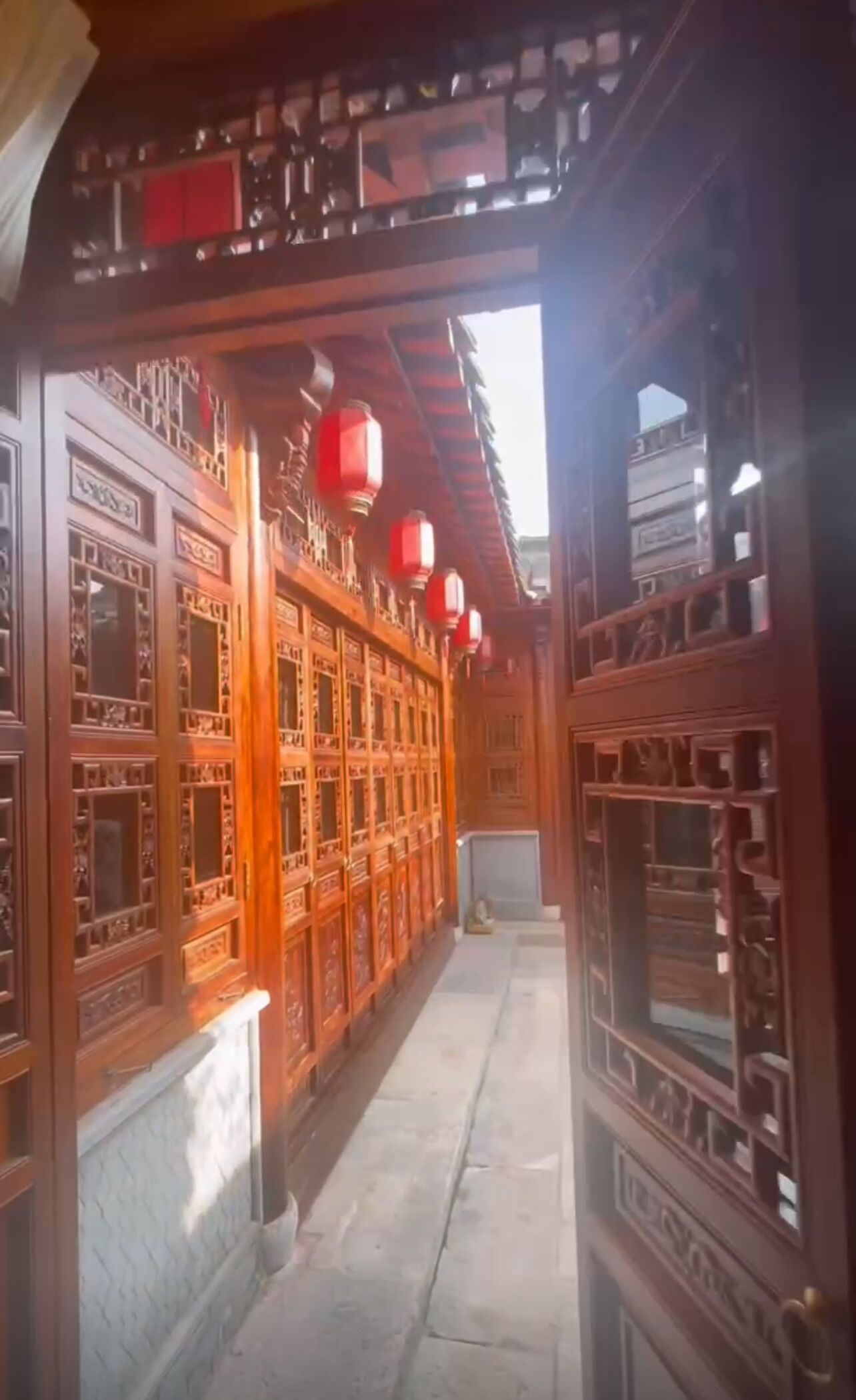 Un pasillo con puertas y ventanas de madera, faroles rojos y suelo de baldosas.