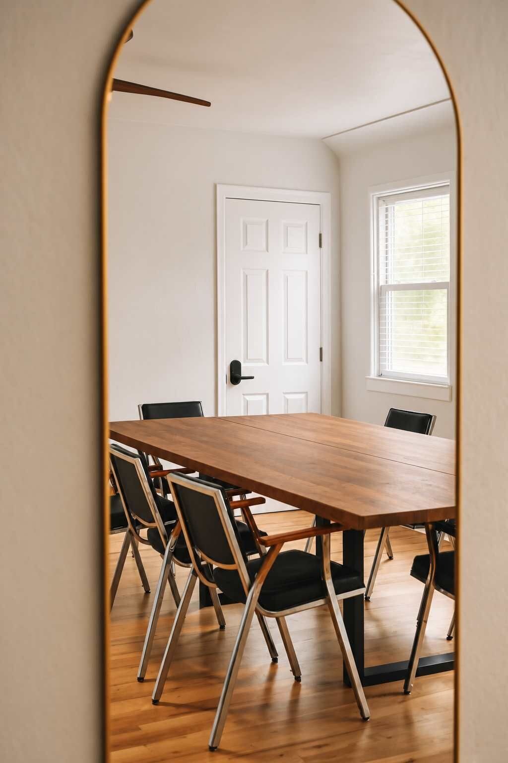 Un salón de conferencias con una mesa de madera y sillas negras, una puerta blanca y un ventanal con persianas.