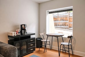 Ein kompakter Wohnbereich mit Küchenzeile, einer Essecke mit zwei Stühlen, einer Couch und einem Fenster mit Blick auf einen Holzzaun.