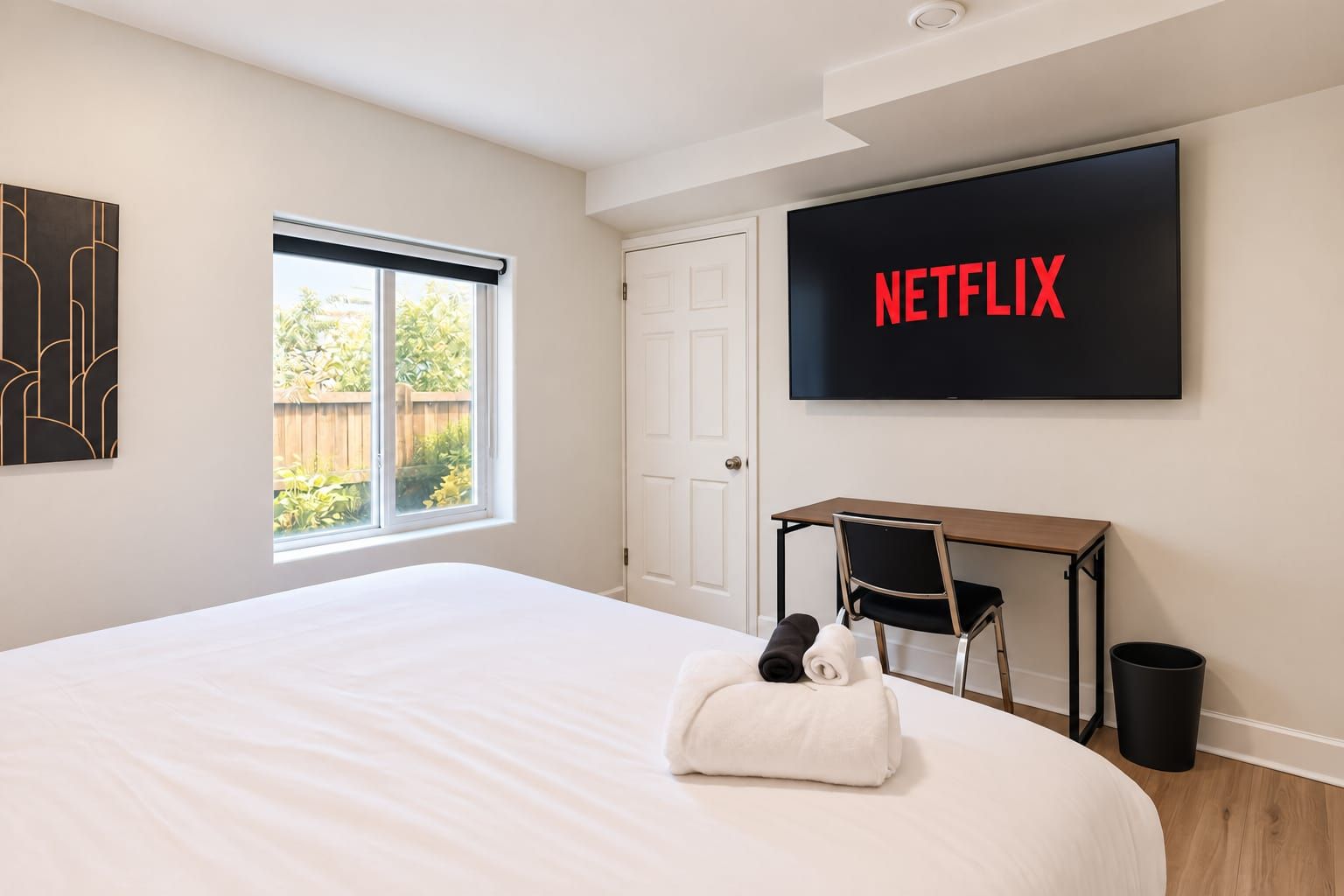 A hotel room with a bed, a desk, a chair, a TV displaying 'NETFLIX,' and a window with a view of greenery.