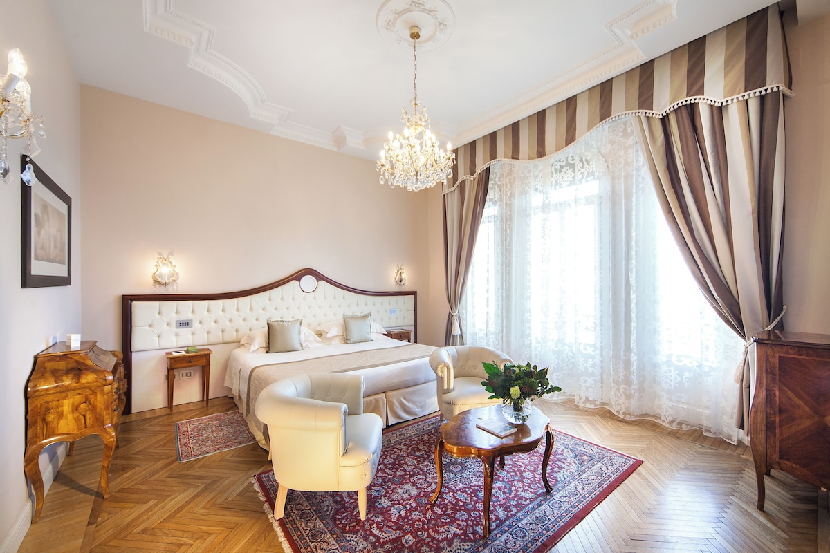 A bedroom with a large bed, a sitting area with a coffee table, a wooden side table, and a chandelier.