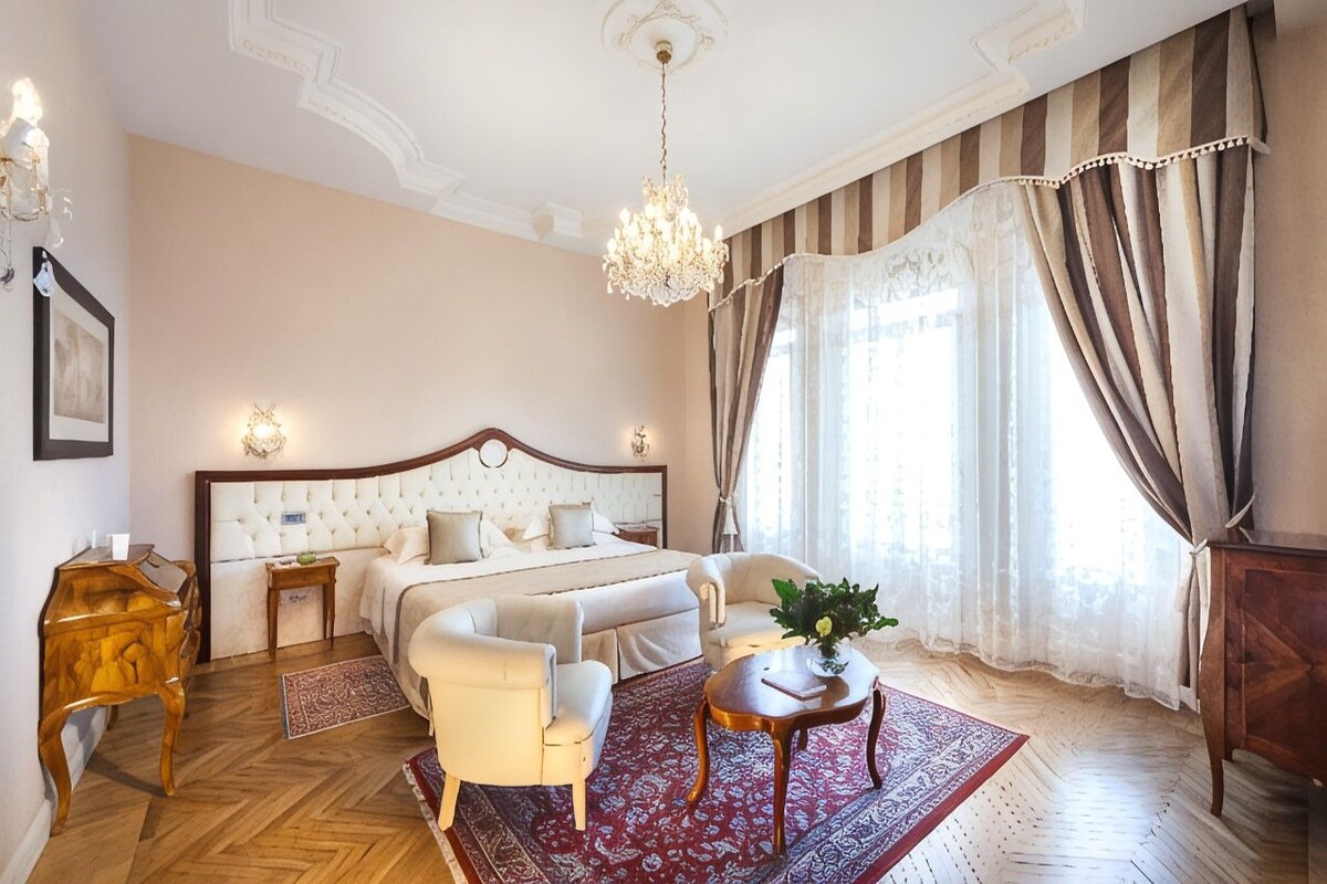 A hotel room with a large bed, a sitting area with a chair and table, a wooden side table, and a chandelier.