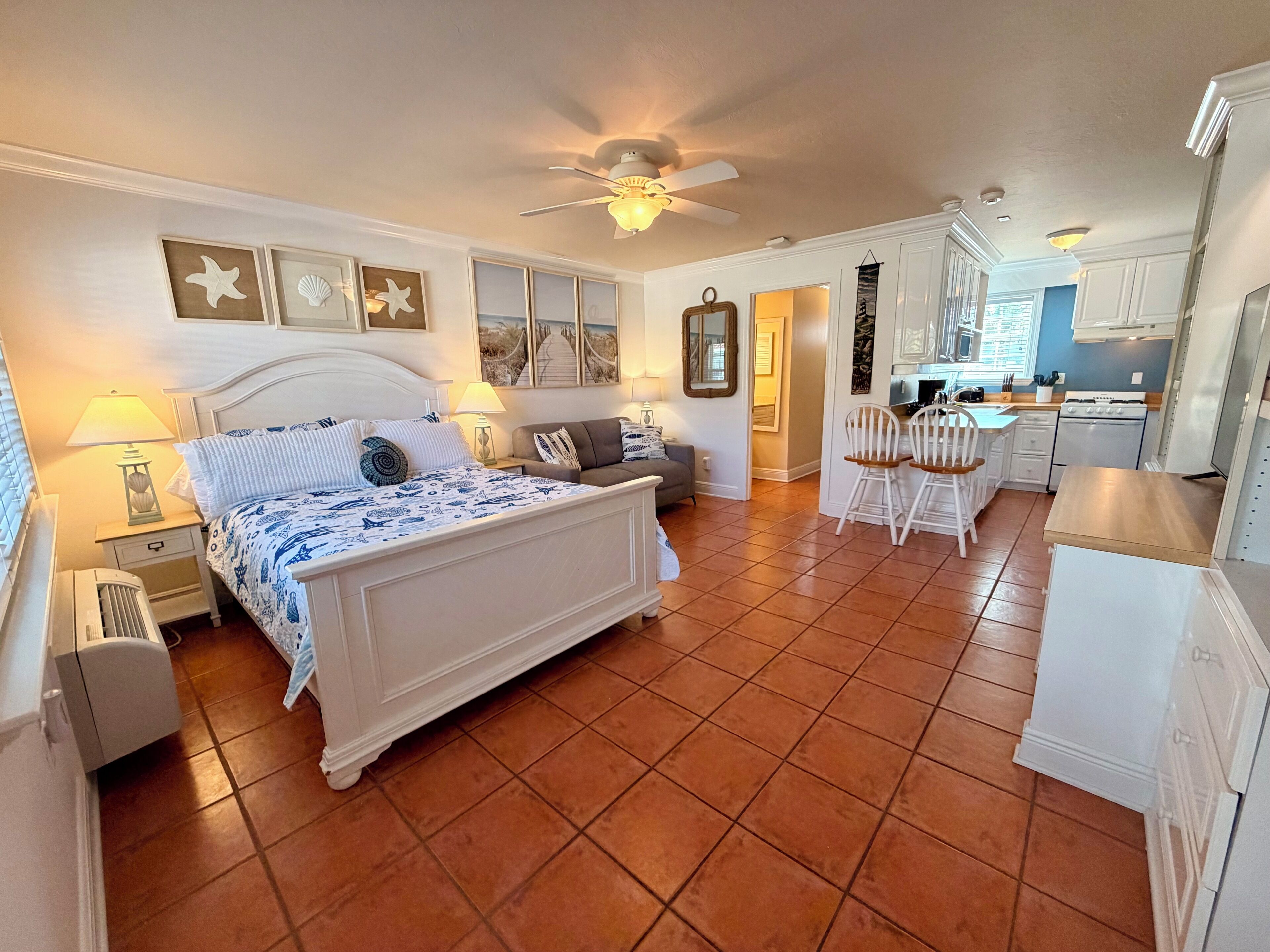 Quarto com cama, mesas de cabeceira, um criado-mudo, ventoinha no teto e vista para a cozinha adjacente.
