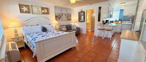 Quarto com cama, mesas de cabeceira, um criado-mudo, ventoinha no teto e vista para a cozinha adjacente.