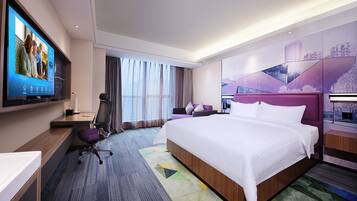 Una habitación de hotel moderna con una cama grande, un escritorio con computadora, un televisor montado en la pared y un ventanal con cortinas.