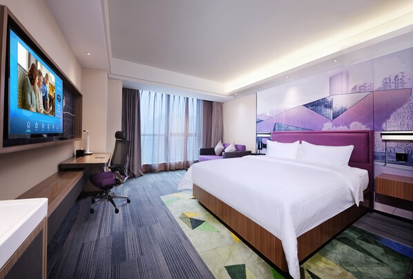 Quarto de hotel moderno com uma cama grande, uma escrivaninha com computador, uma TV na parede e uma janela ampla com cortinas.