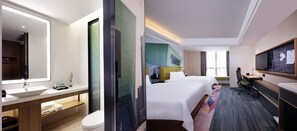 A hotel room with a large bed, a desk with a chair, a television, a bathroom with a sink and mirror, and a window with curtains.
