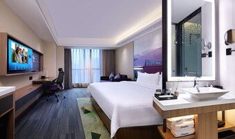 Una habitación de hotel moderna con una cama grande, un escritorio con computadora, televisión y un baño con espejo grande y lavamanos.