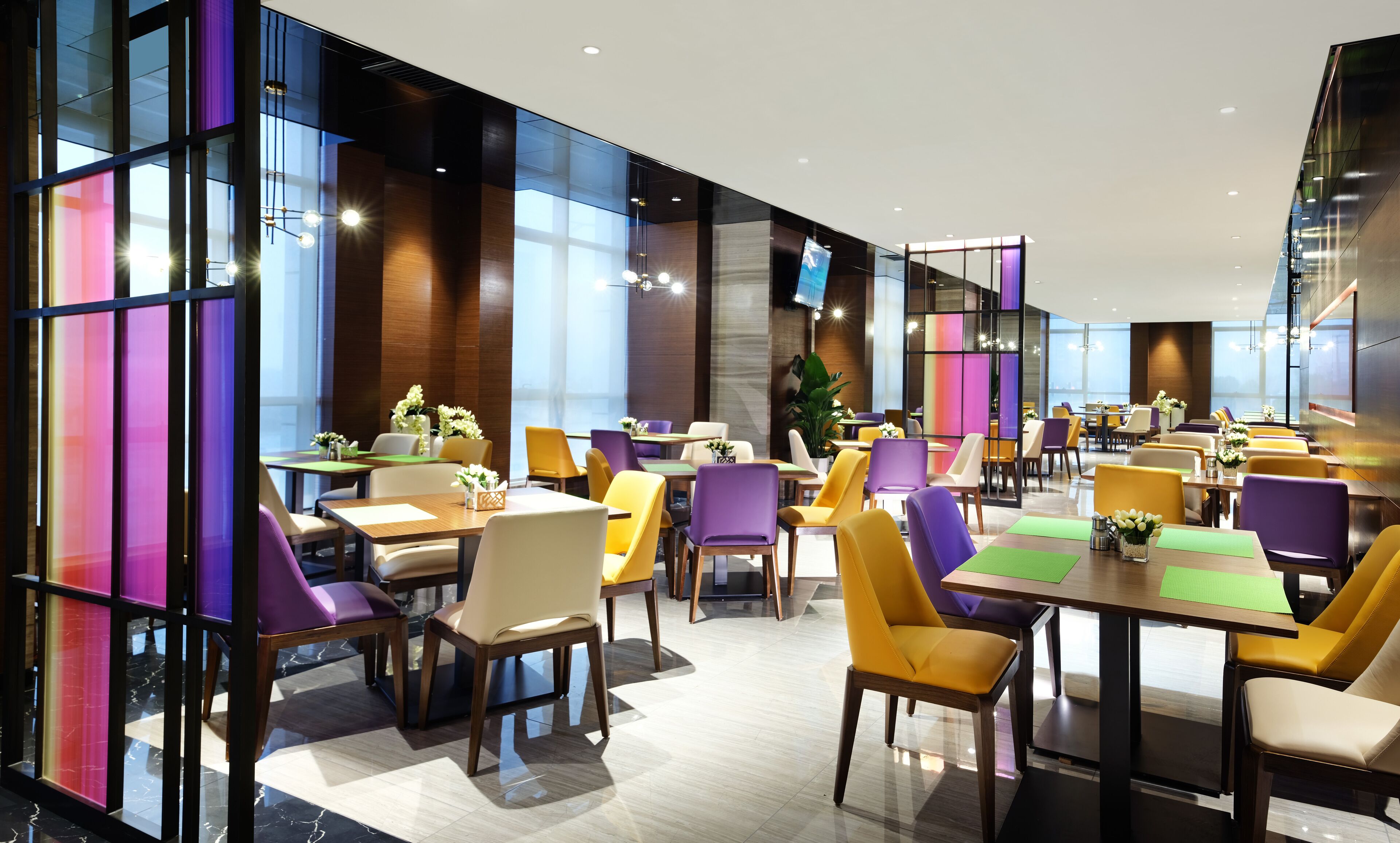 A modern restaurant with multiple dining areas, each featuring a mix of yellow and purple chairs, wooden tables, and glass partitions.