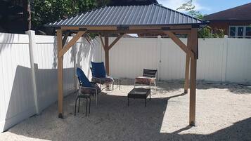 Un área de estar al aire libre con techo, una pérgola, dos sillas, una mesa y una valla blanca al fondo.