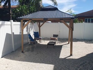 Un área de estar al aire libre con techo, una pérgola, dos sillas, una mesa y una valla blanca al fondo.