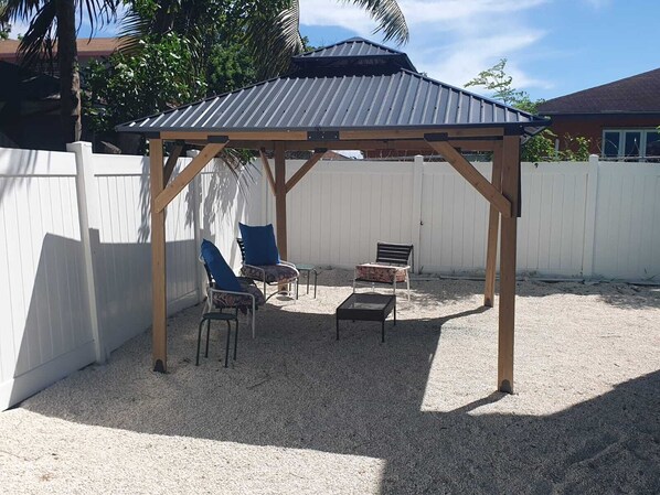A covered outdoor seating area with a pergola structure, two chairs, a table, and a white fence in the background.