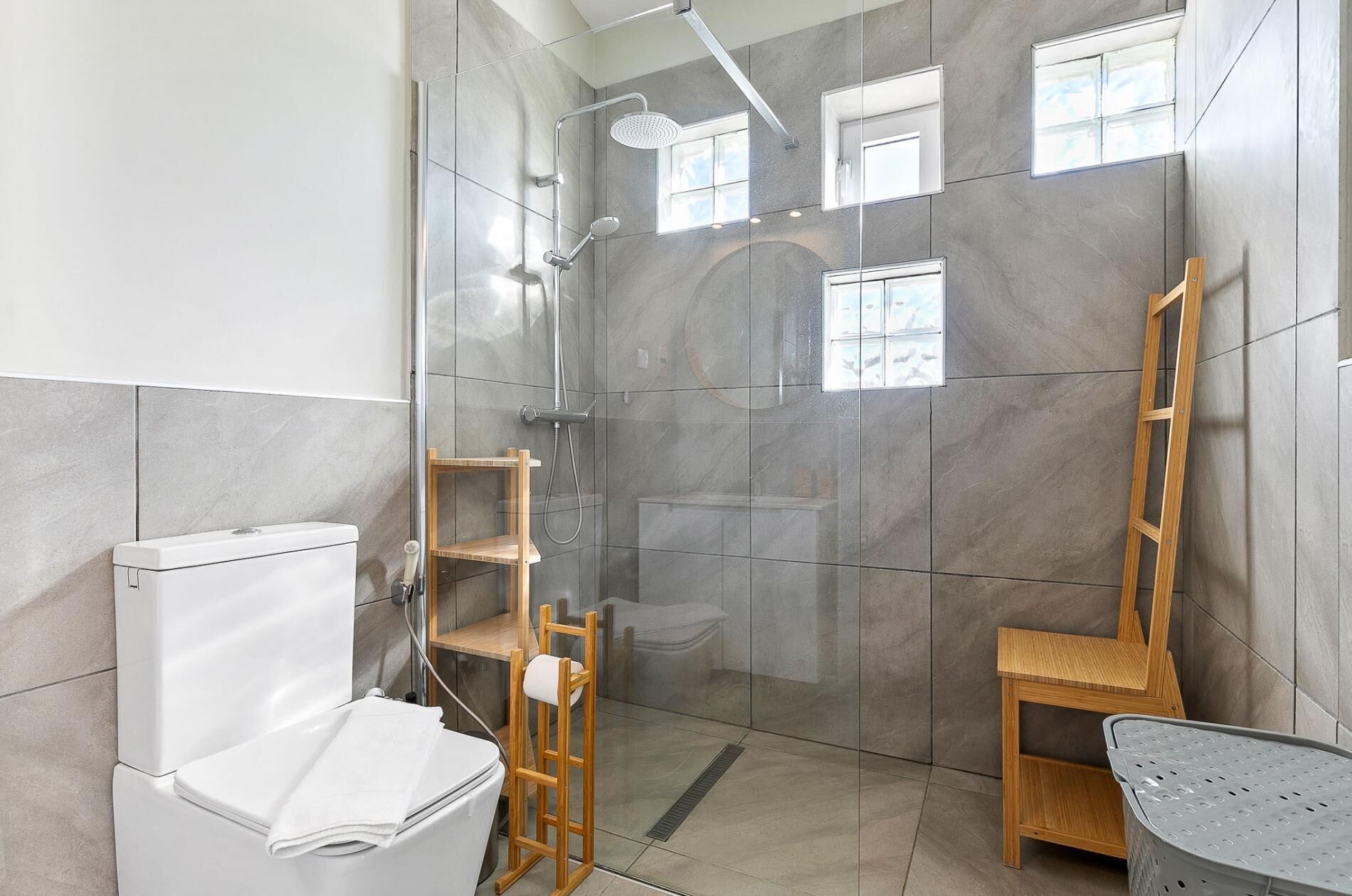 A modern bathroom with a glass-enclosed shower, a toilet, a wooden shelf, and a laundry basket.