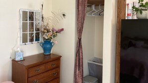 A bedroom with a wooden dresser, a blue vase with flowers, a mirror, a closet with hanging clothes, and a trash bin. - Cozy 1-bedroom studio barn cottage in tranquil Winters (Winters)