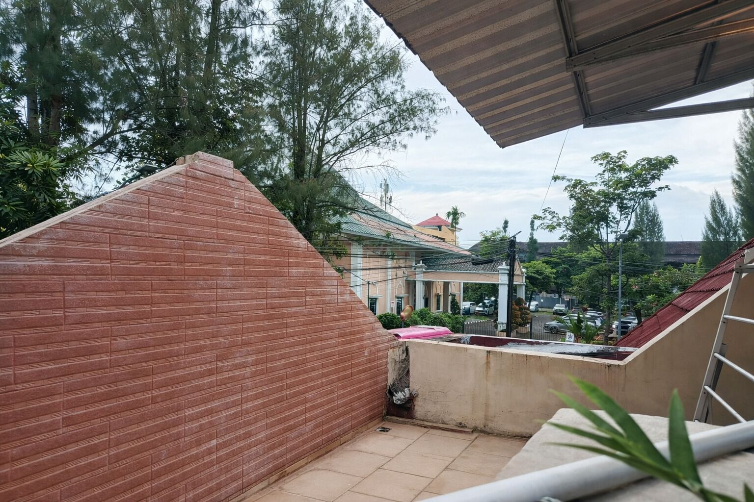 Een overdekt balkon met een bakstenen muur, betegelde vloer en uitzicht op een woonwijk met huizen en bomen.