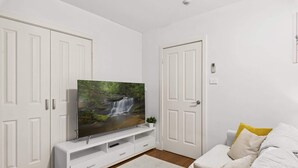 A modern living room with a flat-screen TV on a white entertainment unit, a white sofa with yellow cushions, and a white door with a lock. - Ground Floor One-Bed with Courtyard Access (Strathfield)