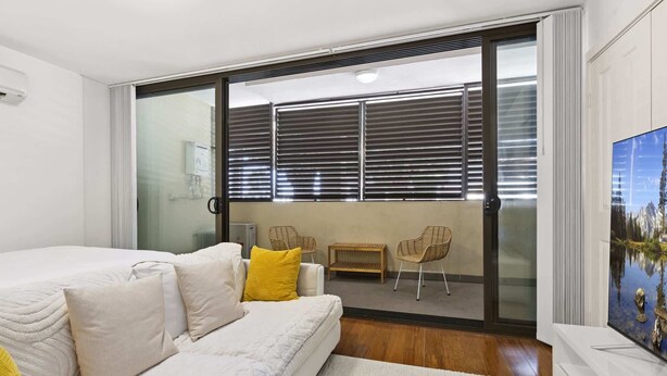 A bedroom with a bed, a sofa, a small table, and a chair. There is a sliding glass door leading to a balcony with a view of the outdoors. - Ground Floor One-Bed with Courtyard Access (Strathfield)