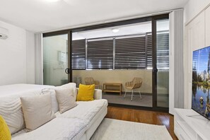 A bedroom with a bed, a sofa, a small table, and a chair. There is a sliding glass door leading to a balcony with a view of the outdoors.