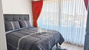 A neatly made bed with a grey quilt and pillows, a grey upholstered headboard, and a view of buildings through sheer curtains. - Moderno impecable Cercano a terminales (Temuco)