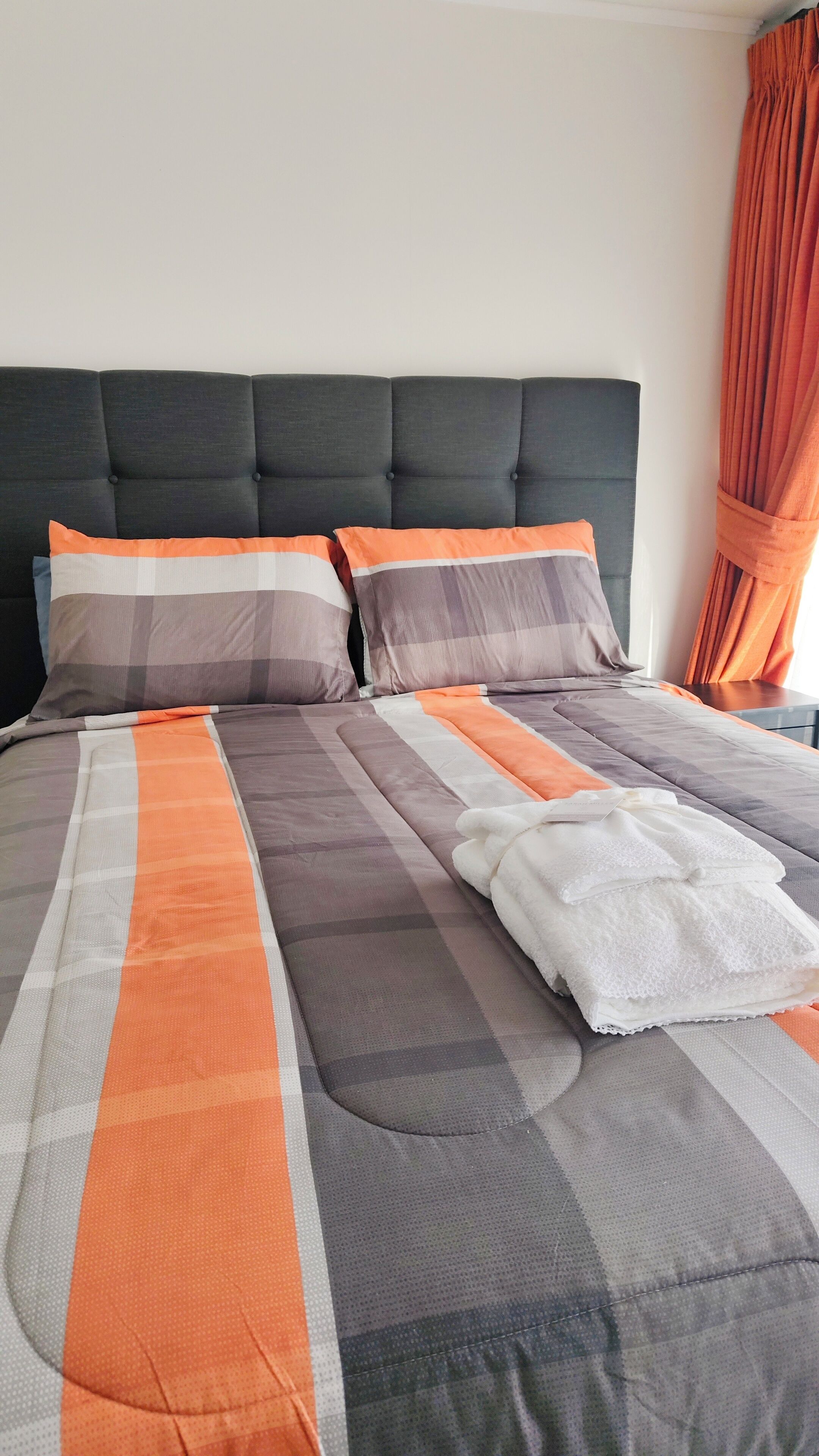 A neatly made bed with a grey and orange striped bedspread, two pillows with similar patterns, and a dark upholstered headboard.