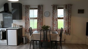 A compact kitchen with a stove, sink, and cabinets, a dining area with a table and chairs, and a view of a grassy field through patterned curtains. - Binnian View Apartment - Sleeps 4 (Newry)