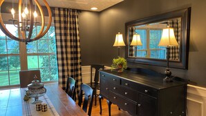 A dining room with a wooden table, chairs, a black dresser, a chandelier, and a window with checkered curtains. - Large Home with Entertainment Options Galore (Elizabethtown)