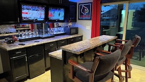 A modern kitchen with a bar area, two TVs mounted above the counter, and a glass door leading outside. - Large Home with Entertainment Options Galore (Elizabethtown)