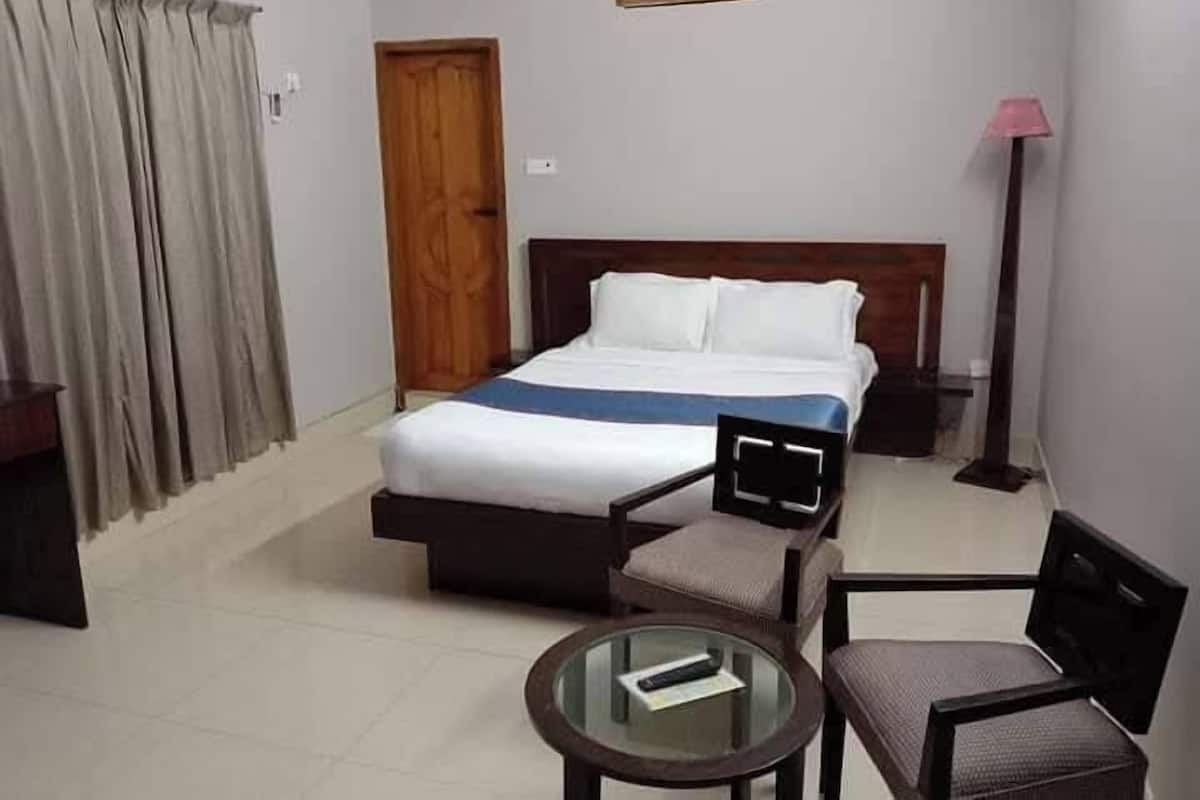A hotel room with a bed, two chairs, a small table, a lamp, and a door.