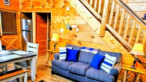 Ein gemütliches Blockhauss-Interieur mit einer Holztreppe, einer Couch mit blau-weißen Kissen, einem Esstisch mit Stühlen und einem Kühlschrank.