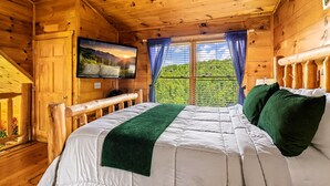 Ein Zimmer in einer Holzhütte mit einem Bett, einem an der Wand befestigten Fernseher und einem Fenster mit Vorhängen.