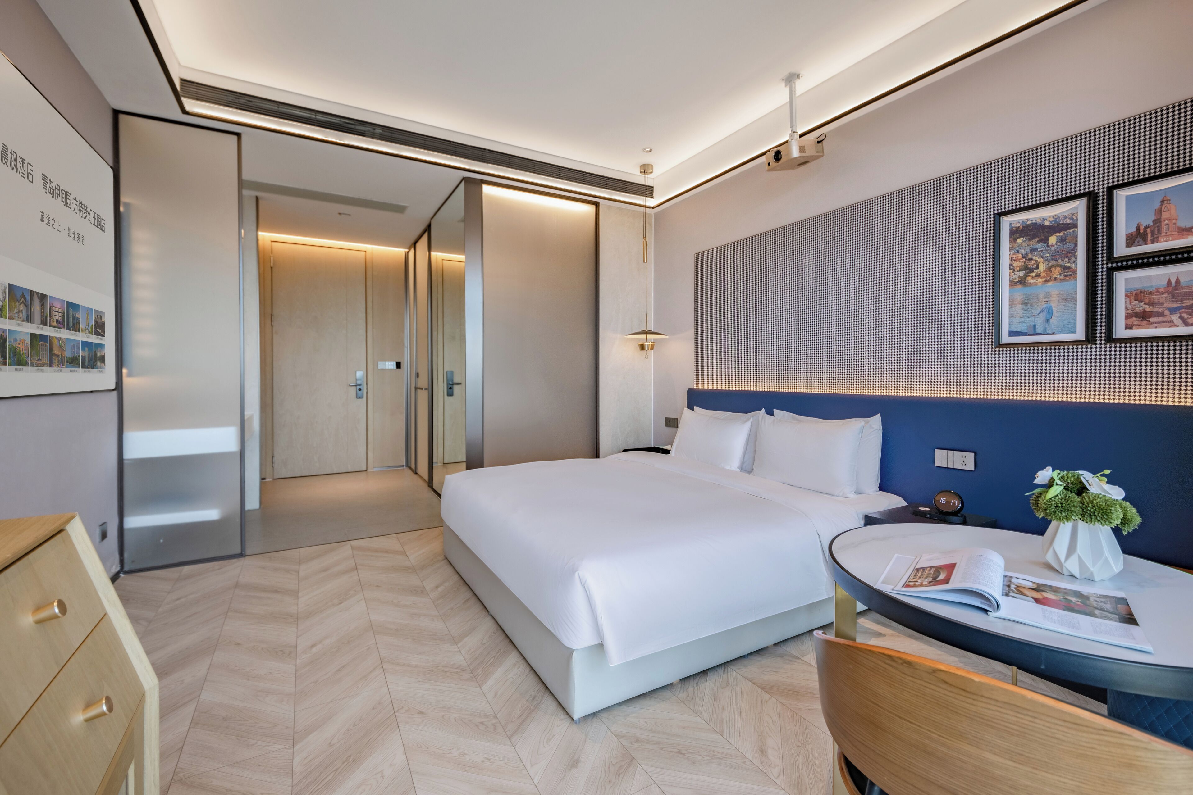 A modern hotel room with a large bed, a bedside table with a magazine, a small round table with a vase of flowers, and a wall-mounted artwork.