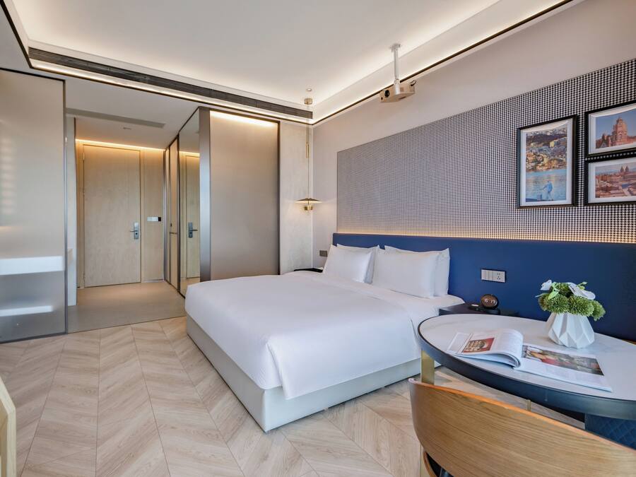 A modern hotel room with a large bed, a bedside table with a magazine, a small round table with a vase of flowers, and a wall-mounted artwork.