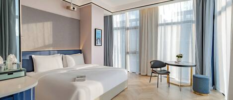 A modern hotel room with a large bed, a desk with a chair, a small table with a vase, and a large window with curtains.