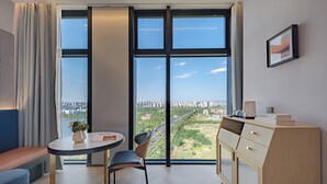 A modern hotel room with a large window offering a city view, a small round table with a chair, a bench, and a wooden cabinet with drawers. - Chenfeng Hotel (Qingdao)